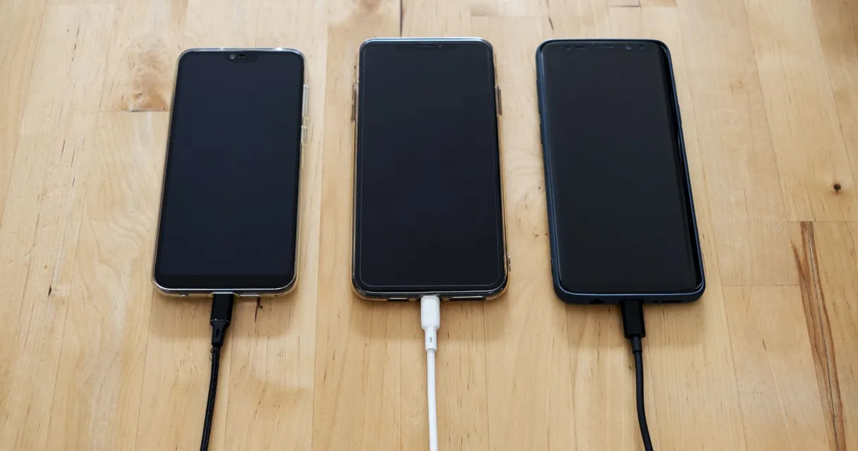 Três celulares ligados ao carregador sobre uma mesa de madeira, ilustrando como economizar bateria do celular